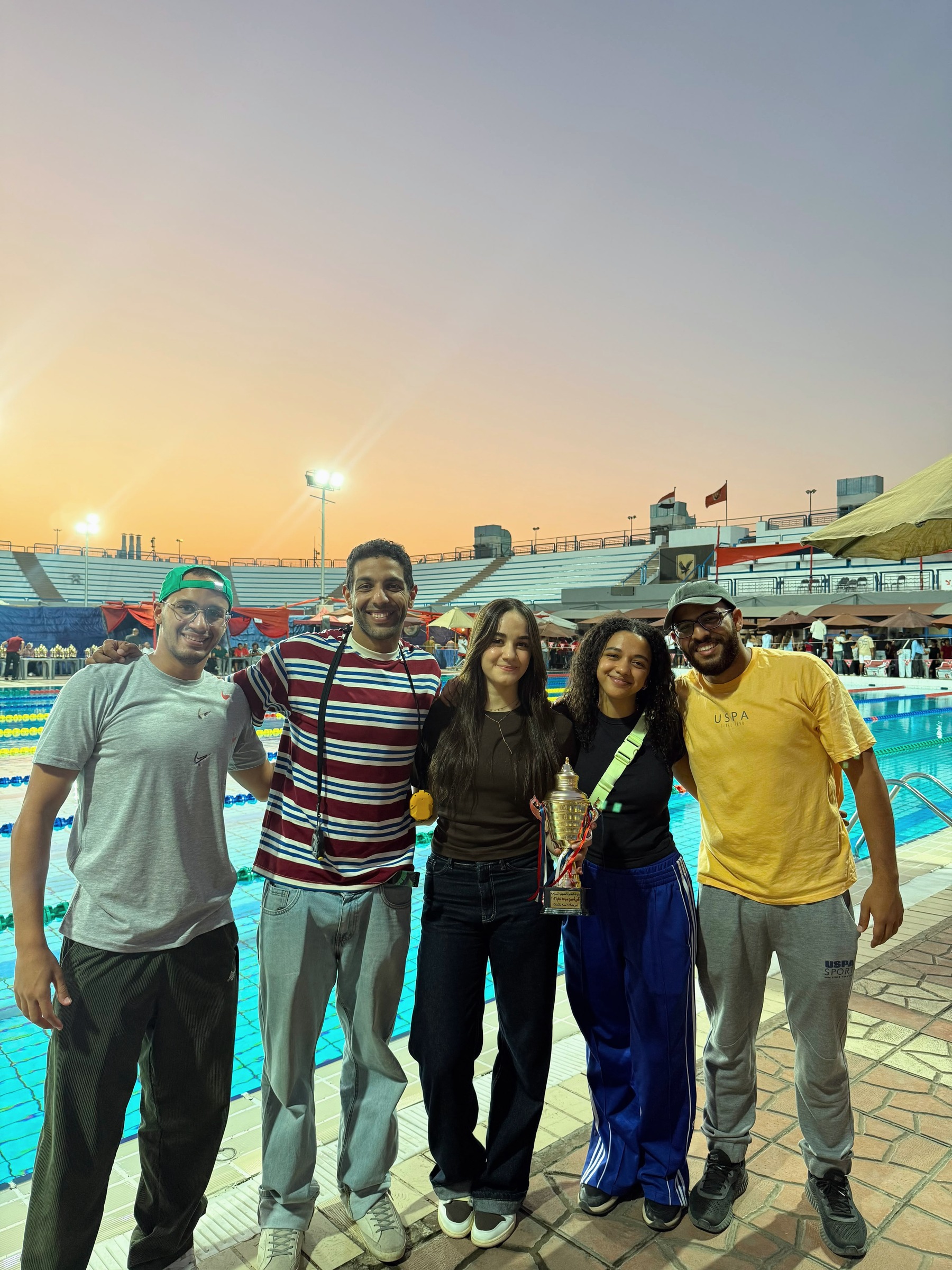 Coaches Amr Fathy, Omar Elewa, Farah Elewa and Ali Hany with a SWEEP swimmer and the meet trophy, by the pool at sunset